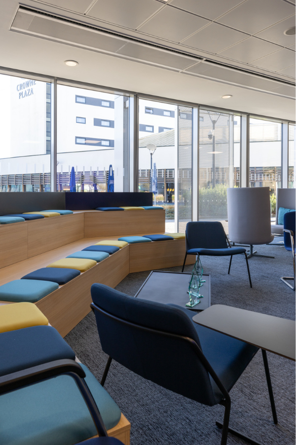 An open plan meeting area with tiered seating and poseur height collaboration tables. The desk area can be seen behind.